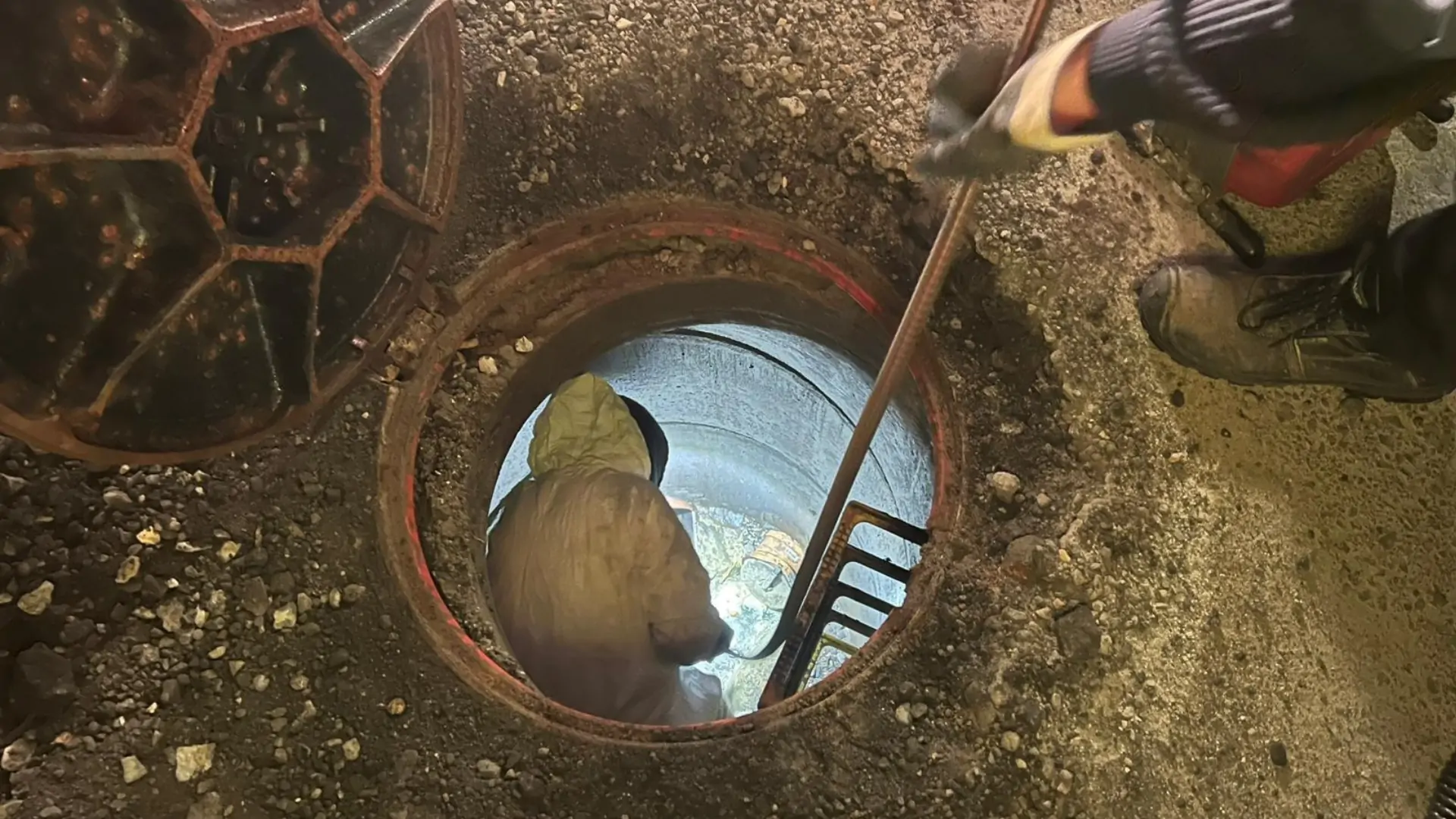 Otwarty właz od kanalizacji i wchodząca do niego po drabinie osoba.